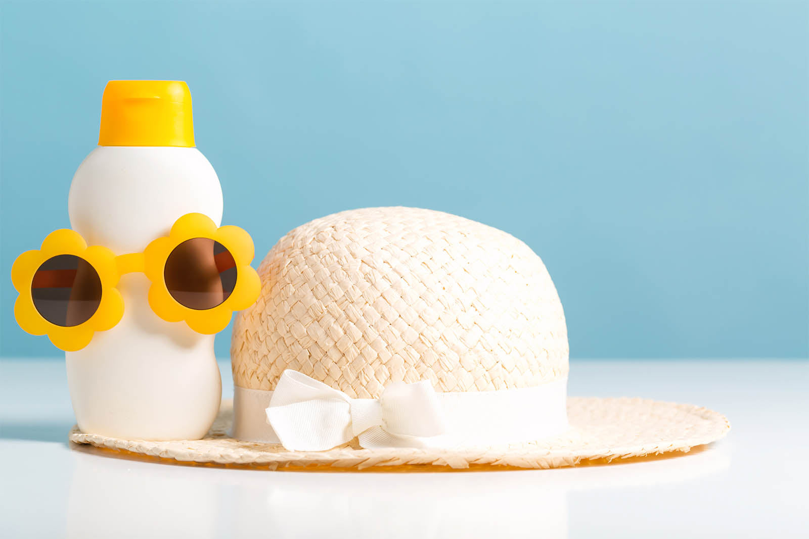 A white bottle of sun block wearing a pair of yellow sunglasses. The bottle is resting on the brim of a pink ladies sun hat with a white bow around the base.